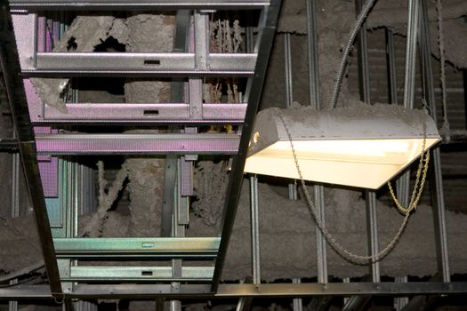 Industrial Rafters Construction Dust is a capture of an exposed area. There is a mix of insulation and dust visible next to a light that is creating a rainbow effect on the structure that looks like a ladder.
