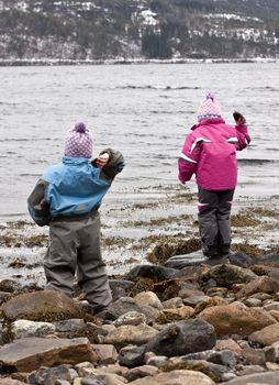 Two children standing on a rocky beach, throwing stones into the sea