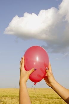 Woman hands holding red balloon. Agricultural field and sky with clouds in background.