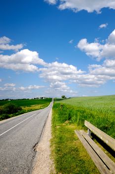 Road with surrounding green fields. The sky is blue with white clouds. On the left is a bench.