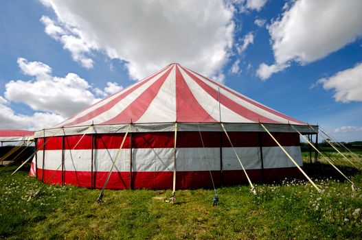 A red and white striped circus tent in green nature. The sky is blue with white cumulus clouds