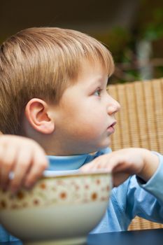 Cute little blond boy sitting at the table, holding a bowl and looking away