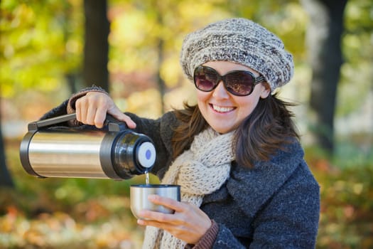 Young pretty woman with glasses and cap drinks tea in autumn park