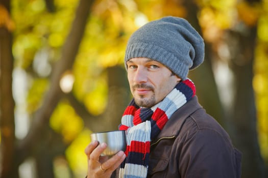 Young happy man with scarf and hat drinks tea in autumn park