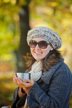 Young pretty woman with glasses and cap drinks tea in autumn park. Vertical view