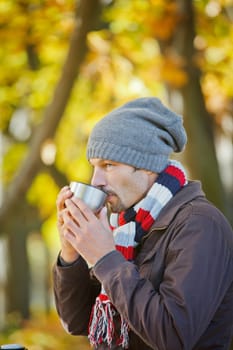 Young happy man with scarf and hat drinks tea in autumn park. Vertical view