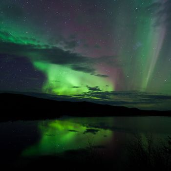 Night Sky Stars, clouds and Northern Lights mirrored on calm lake in Yukon, Territory, Canada.