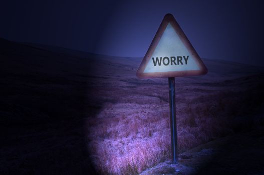Night shot of a single road warning sign with the word 'worry' illuminated by car headlights with subtle remote and dark landscape behind.