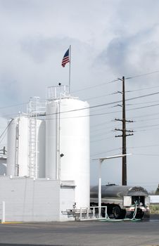 Vertical image of a tanker filling his truck from tall white silos with an American flag flying from the top.