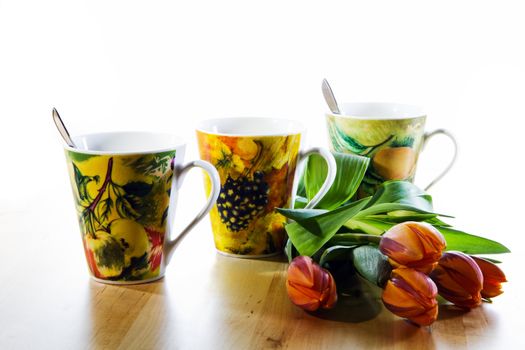 Colorful still-life with three decorated mugs, orange tulips and white background on wooden table