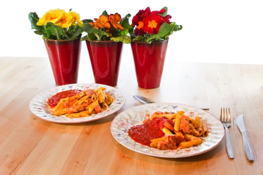 Colorful still-life- two plates penne macaroni with spicy ham and tomato-basil sauce - three primroses in red pots as a spring decoration on wooden table