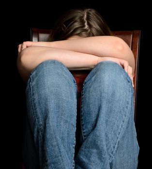 A sad girl holding her head in her arms, while sitting on a chair, isolated on black.
