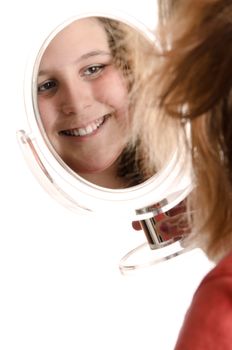 A smiling preteen girl is looking at herself in a small mirror, isolated against a white background.
