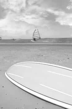 surfboard lying on the beach as surfers windsurf in the maharees in county kerry ireland during a storm in black and white