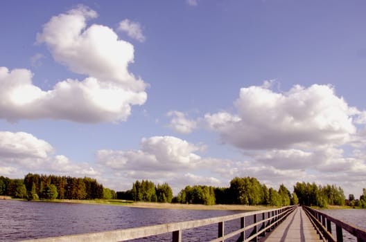 Long wooden footbridge with handrails over the lake. Forest in the distance and the cloudy sky.