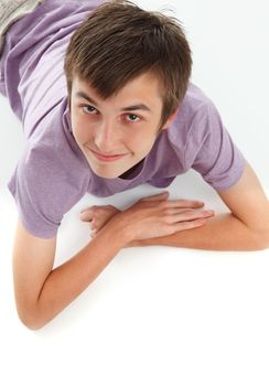 A smiling relaxed boy in a purple t-shirt is resting lying on stomach and looking up.  White background.