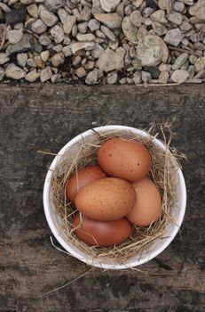 A bowl of freshly laid organic brown eggs, set in a white ceramic bowl with straw. Set on a background of wood and stone gravel. Set on a portrait format with copy space available.