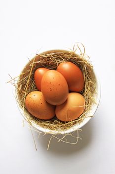 Five freshly laid organic brown eggs, set in a white ceramic bowl with straw. Set on an isolated white background.