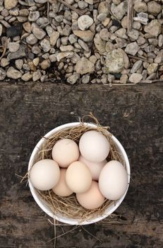 A bowl of eight freshly laid organic white eggs, set in a white ceramic bowl with straw. Set on a background of wood and stone gravel. Set on a portrait format with copy space available.