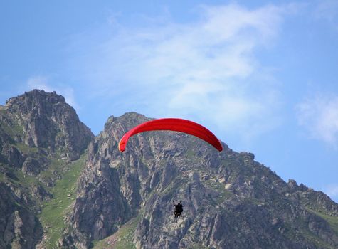 Red paraglider flying in the sky next to a rocky mountain