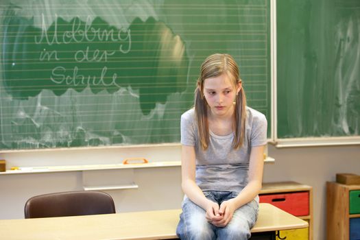 A sad girl sitting in the classroom at school in front of a blackboard with the words "bullying at school"

