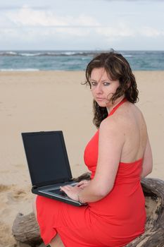 Lady in red dress typing on a laptop at the beach