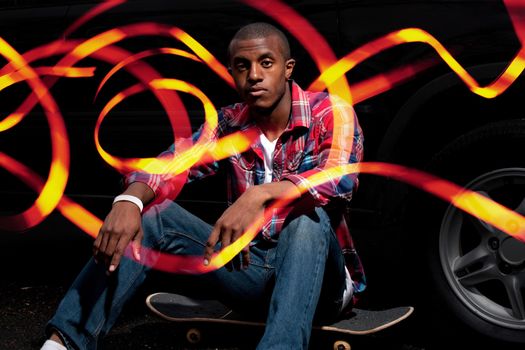 A young man hanging out seated on his skateboard with abstract light trails glowing around him and through his fingers.