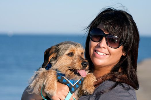 A young woman in her 20s by the sea shore holds a cute mixed breed beagle yorkshire terrier dog also called a Borkie.