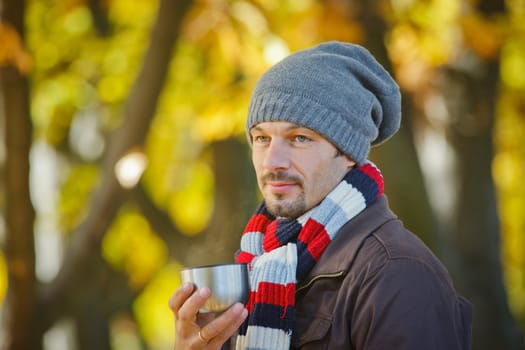 Young happy man with scarf and hat drinks tea in autumn park