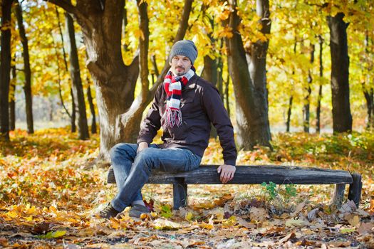 Young attractive man with scarf and hat resting on a bench in autumn park