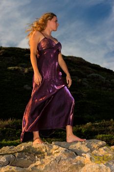 Girl standing on a rock in a purple dress and her hair blowing in the wind