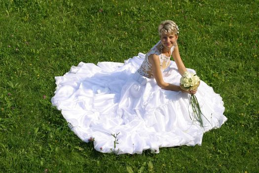 The beautiful bride with a wedding bouquet sits on a grass
