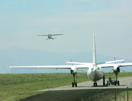 One aircraft takes off while another sits on the runway as a mechanic works on it.