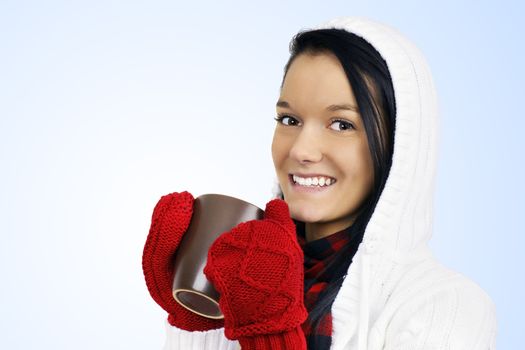 Winter time concept: cute smiling friendly natural young woman with red mittens and white hoodie drinking hot chocolate, coffee or tea in a brown mug over light blue gradient background.