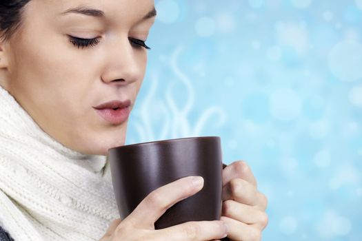 Winter time concept: friendly natural young woman with white wool scarf blowing on her hot chocolate, coffee or tea in a brown mug over light blue fun background.