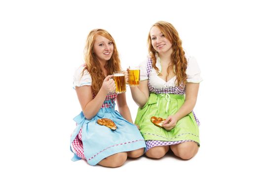 two bavarian girls with pretzels and beer kneeling on floor on white background