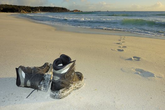 A pair of boots on a sandy beach in the early light of dawn Footprints lead into the sea but do not come out again. Space for copy on the sand.