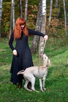 A girl in a black dress and white saliki pup in a forest 
