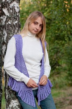 A girl in a lilac vest and white polo-neck sweater and jeans in a forest
