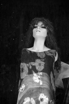 Plastic mannequin doll behind stained window of abandoned store. Black and white.