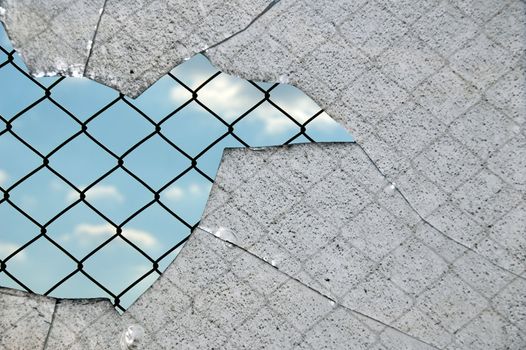 Broken glass fragments and chain link wired fence pattern against blue sky.