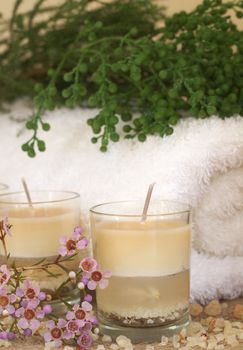 Relaxing spa scene with a white rolled up towel, pink flowers and green filler, beautiful handmade candles and bath salts
