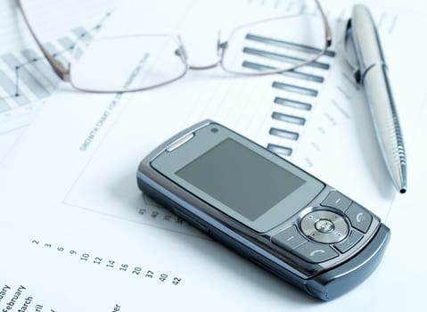 Close-up of cellphone with modern silver pen and reading glasses on top of potential growth charts and graphs. Blue tone image