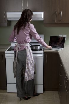 Young woman with apron, standing in front of the stove, sturring a pot and working on a laptop