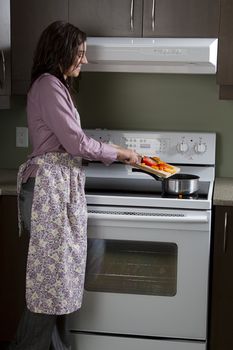 Young woman with apron, standing in front of the stove, putting cut vegetable in a pot