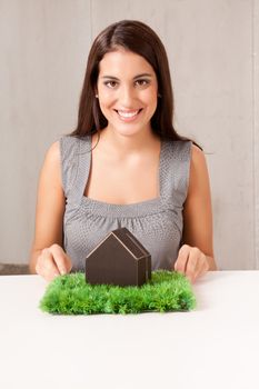 A woman holding a model house which is sitting on a turf of grass