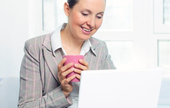 Attractive smiling young business woman with cup using laptop at work desk