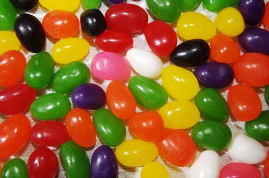 Colorful, tasty jelly beans spread out over a flat surface. The candy is viewed from directly above, and fills the frame entirely with orange, yellow, white, black, pink, green and red.