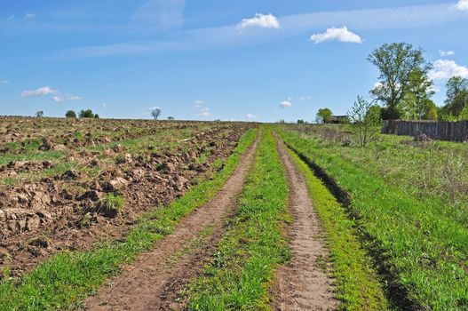 Country dirt road along the ploughed field at village outskirts, spring sunny day