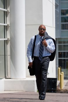 A confident business man carries his suit jacket over his shoulder while walking down the sidewalk in the city.